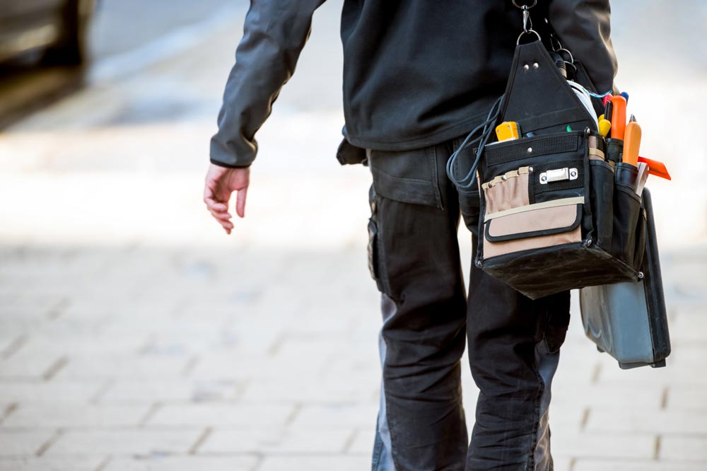 Elektriker i fritidskläder går på gatan med välfyllt verktygsbälte och verktygsväska i handen